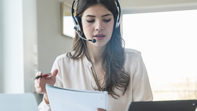 Woman with phone headpiece looking at papers and her laptop.
