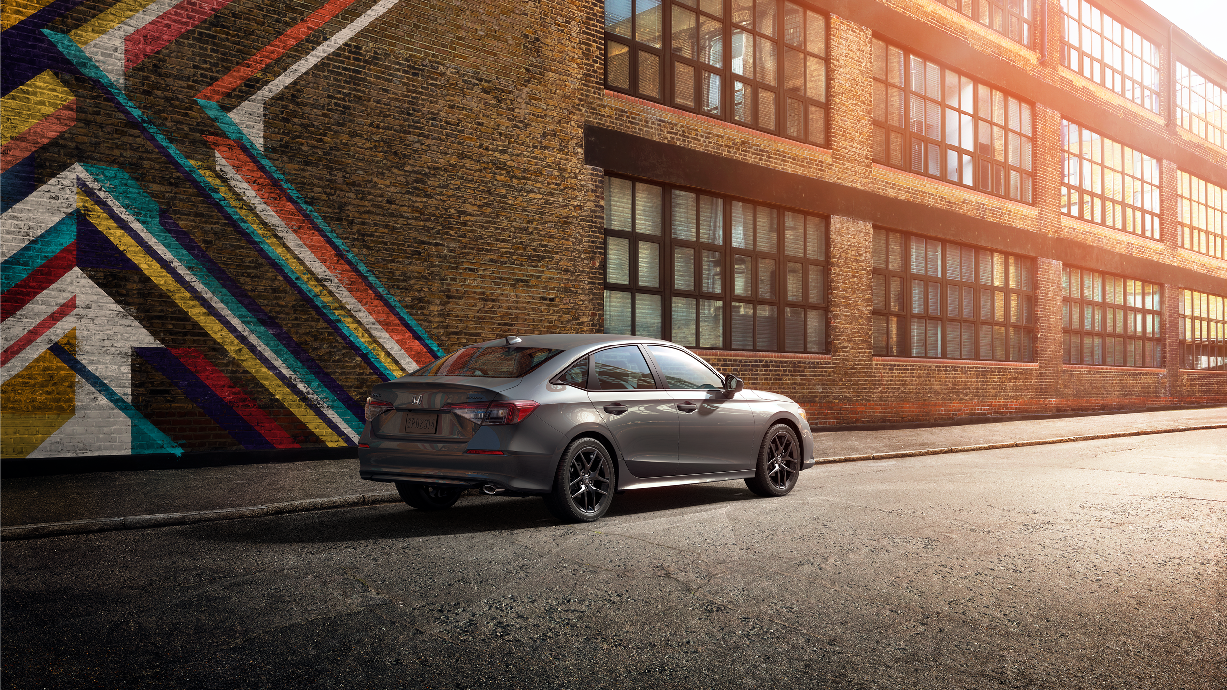 3/4 rear view of grey Civic Sedan in an old factory district parked next to a graffitied brick wall.