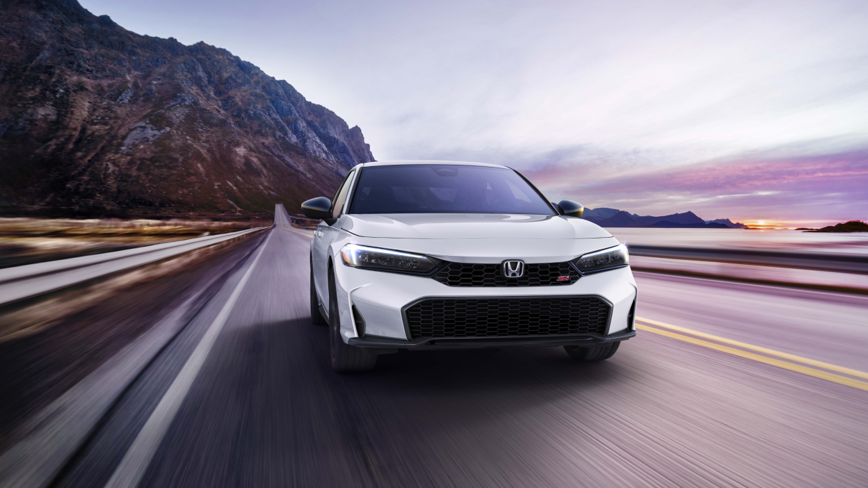 Front view of white Civic Si driving toward camera on a mountainous oceanside highway.