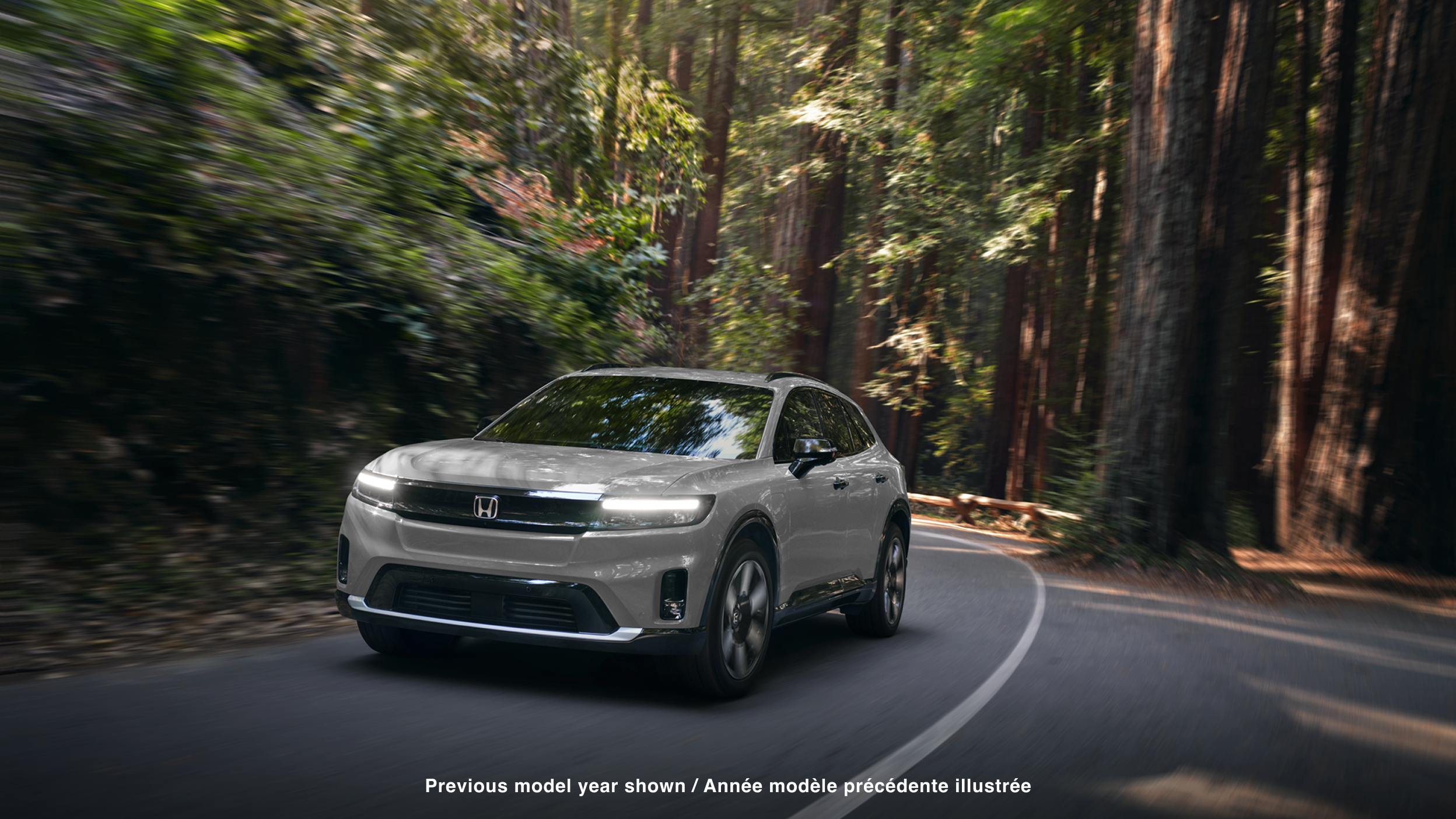 3/4 front view of white Prologue taking a turn on a paved road in a temperate forest.