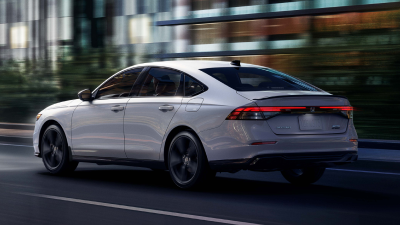 3/4 rear view of white Accord driving on city side highway past buildings at night. 