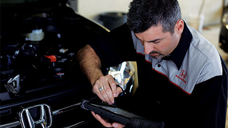 Honda technician kneeling in front of the open hood of a black Honda, performing an inspection and reviewing information on a black tablet. // Un technicien Honda à genoux devant le capot ouvert d'une Honda noire, effectuant une inspection et examinant des informations sur une tablette noire.